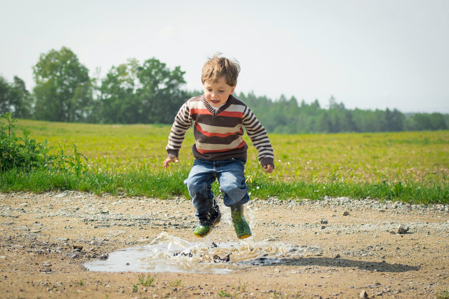 Child in a striped sweater and rain boots jumping into a puddle outdoors.